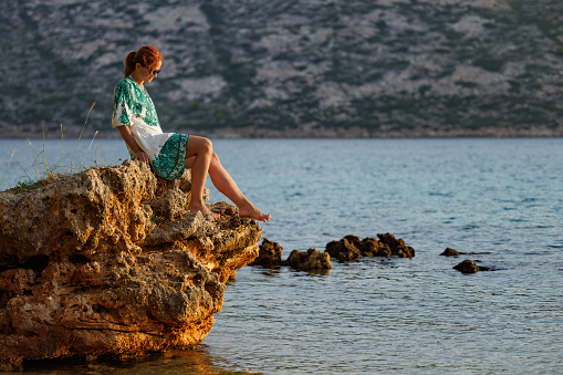 Beautiful redhaired woman in summer dress sitting on a boulder by the sea, enjoying solo vacation on the island