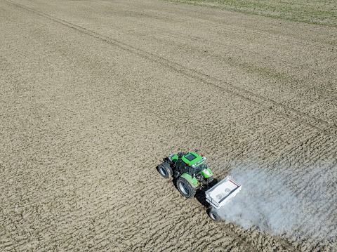 Tractor spreading agricultural lime as a soil additive in a field seen from above. There is dust coming from the spreader.