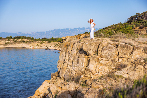 Beautiful woman in white dress and sunhat, standing on a cliff by the sea, enjoying solo vacation on the island
