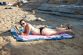 Teenage girl in bikini wearing sunglasses and smiling, lying on beach towel on the sand