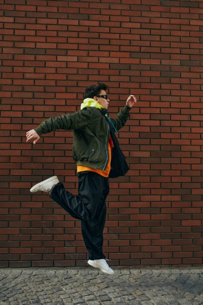 istock Trendy dressed young guy in sporting jacket and sunglasses jumping in motion against brick backdrop. 2149967774