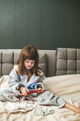 A cute little girl in a bathrobe sitting comfortably on the bed, completely absorbed in the digital experience, lost in the smartphone. Concept: kids and smartphones