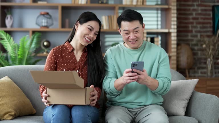 Joyful asian family couple customer opens package at home. Happy wife and husband buyer unpacking purchase sitting on sofa