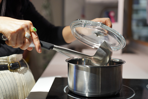 Selective focus, Hands of woman cooking delicious meal in pot on electric stove in kitchen, she holding glass pot lid and ladle in hand.