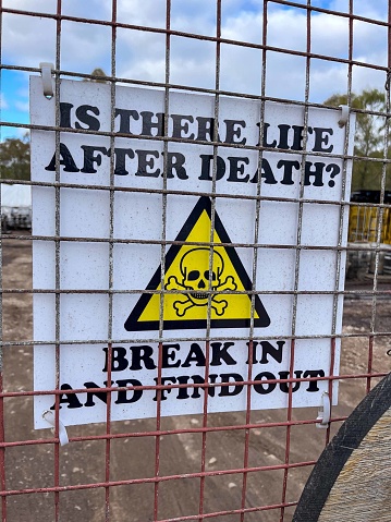 Harsh signage fixed to a yard wire gate, warning potential intruders not to try and break in, by asking the question - Is there life after death? To add weight to the dark humour message a skull and crossbones illustration has been added in a yellow triangular sign.