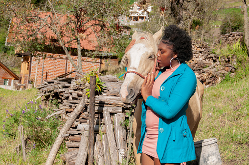 A woman in a blue jacket tenderly hugs a white horse near a wooden fence with rustic houses in the background.