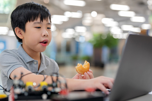 Boy sits in a classroom, focused and concentrated, as he writes code on a computer while enjoying donuts. Multi-tasking approach to studying, combining learning with a moment of relaxation and enjoyment. Student's curiosity, intelligence, and creativity in computer programming and technology.