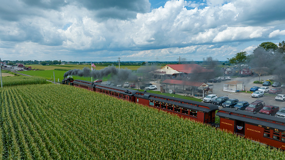 Ronks, Pennsylvania, August 15, 2023 - Clouds gather above as a historic steam train runs parallel to lush cornfields, offering a journey through time by the rural landscape.