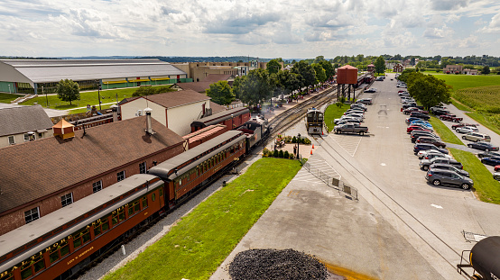 Strasburg, Pennsylvania, August 15, 2023 - A vintage steam train is poised for departure at a charming railway station, evoking a bygone era against a modern-day backdrop of cars and greenery.