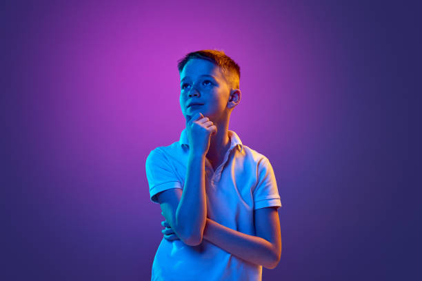 istock Little boy, school-boy with thoughtful expression looking away in neon light against purple gradient background. 2149986611
