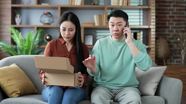 Unhappy asian family couple arguing on phone after receiving a parcel with wrong product at home.