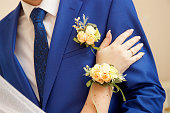 The hand of the bride with a boutonniere rests on the forearm of the groom in a blue suit with a boutonniere and tie