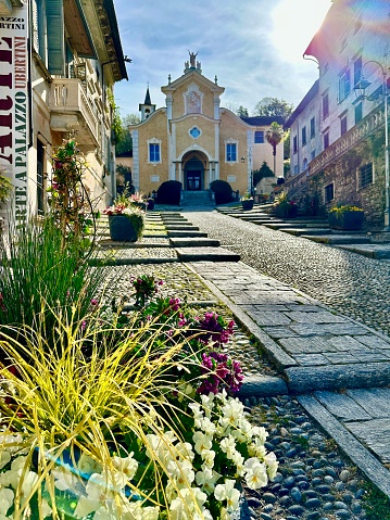 The photo was taken on a spring day at Orta San Giulio, at the edge of Lake Orta (Piedmont). It shows the Parish church of Santa Maria Assunta, Virgin Mary (XV century) standing at the end of a pebbled uphill road, delimited by big flowers pots.