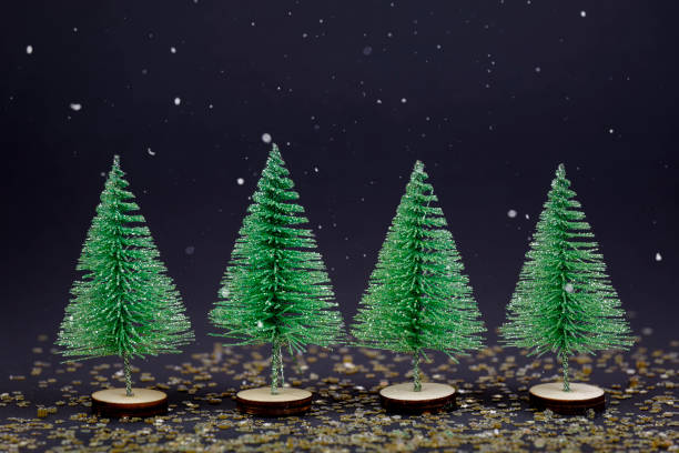 Small green christmas trees in a row on dark background with snowflakes falling down. Winter greeting card background with copy space