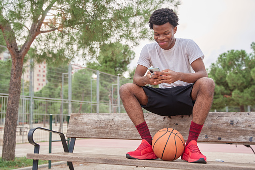 young african-american man using his smart phone sitting on a bench with a basketball