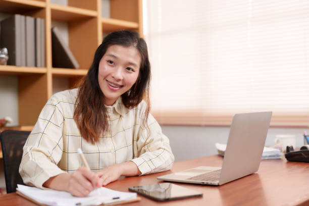 istock Young writing, focused, inhome office setting laptop, indoor daylight illuminating desk. Diligent individual pens notes, technology at hand, serene workspace with natural light, serene ambiance 2149999276