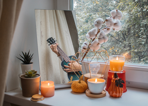 Girl sits in her room on a window sill decorated for the holiday and plays the ukulele. Her reflection is in the mirror next to lighted candles, flowers and pumpkin figurines. Home decoration for holidays, Thanksgiving, Halloween, birthday. Comfort and tranquility
