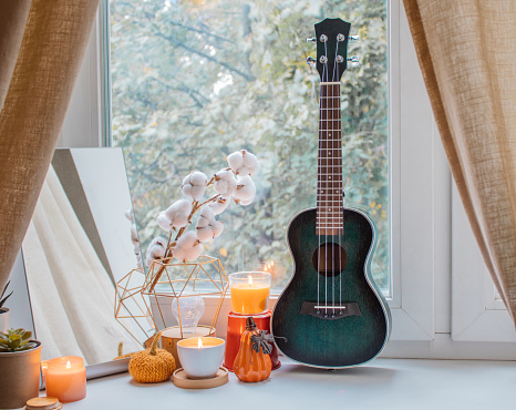 Autumn composition of candles, mirror, flowers, pumpkin figurines and ukulele on the windowsill in girl's room. Home decoration for holidays, Thanksgiving, Halloween, birthday. Comfort and tranquility