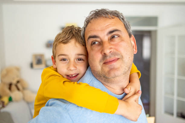 Portrait of an adorable and cute Caucasian boy, embracing his father from behind, while both enjoying time between themself at home