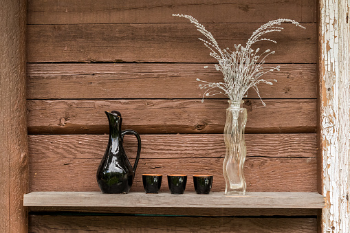 Vase with dry flowers and a jug with small glasses against a brown wooden wall
