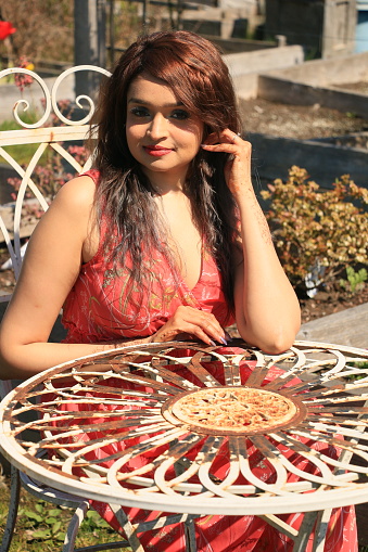 A closeup of a Fijian woman sitting at a rustic iron table and chair in a Community Garden in Spring. She is wearing long, brown straight hair, makeup, and a red sleeveless dress.