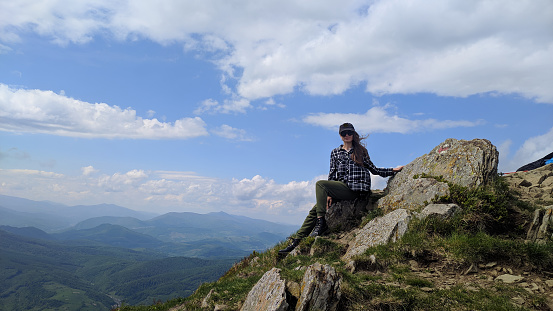 A tourist girl with long hair sits on a stone on a mountain peak, enjoying the beautiful mountain landscape, evoking a feeling of calm and relaxation, active recreation in the fresh air.