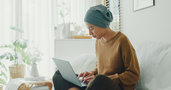 Young Asian woman sick with cancer with headscarf comfortably working on her laptop, seated on a sofa in a well-lit, plant-decorated room. Living with cancer concept.