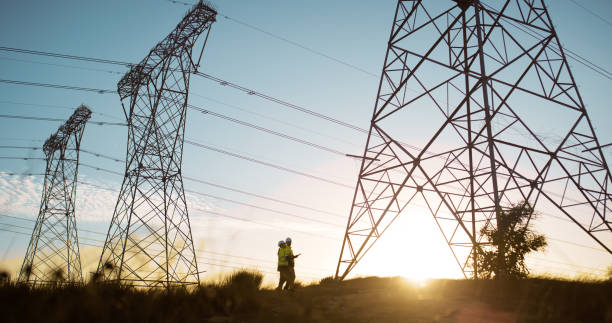 istock People, engineering and transmission tower or lines maintenance for electricity, teamwork or circuit. Sunset, inspection and pylon poles for power supply or steel grid, industrial or infrastructure 2150017022
