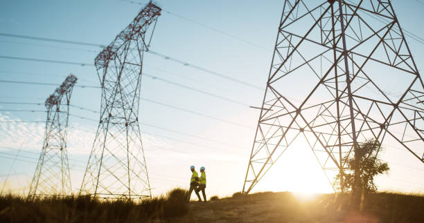 istock People, engineer and transmission tower or industrial with electricity for maintenance, voltage or infrastructure. Sunset, pylon and pole or cables with blue sky for circuit, current or connection 2150017023