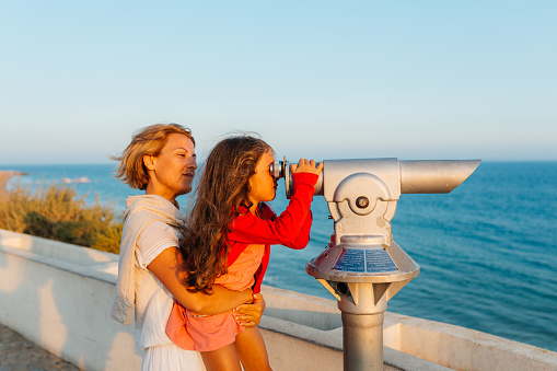 Mother holding her daughter  on a view point and she is  looking through binoculars at the beautiful beach in Albufeira in Algarve Portugal