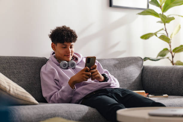teenage boy with smartphone relaxing on living room sofa - tiener fotos stockfoto's en -beelden