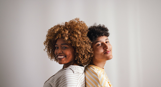 A joyful African American mother and her teenage son smiling as they stand back to back in a studio setting, radiating happiness and warmth.