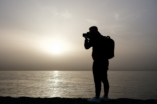 A young man takes a photo on the beach at sunset. Silhouette photo.