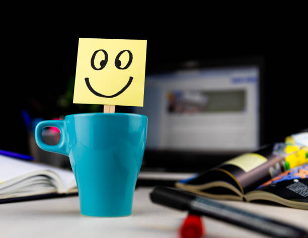 Smiling face drawn on sticky note inside a cup of coffee at the office desk. Every morning wake up with the smile