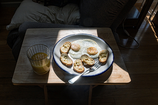 A full breakfast plate with two salt and pepper-seasoned fried eggs and several thin slices of crisply toasted French bateau bread accompanied by half a glass of fresh orange juice. Early morning photo in a living room with strong direct sunlight coming through east facing windows and casting dark shadows.