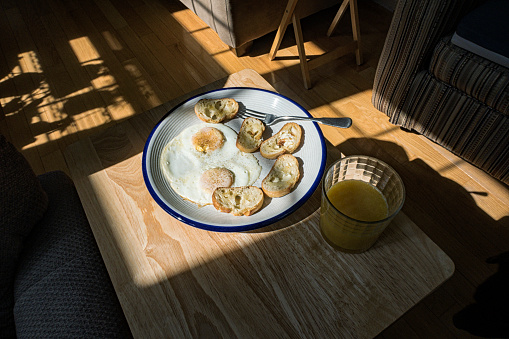 A full breakfast plate with two salt and pepper-seasoned fried eggs and several thin slices of crisply toasted French bateau bread accompanied by half a glass of fresh orange juice. Early morning photo in a living room with strong direct sunlight streaming through east facing windows and casting dark shadows.