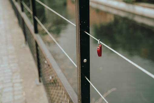 A single red padlock, often symbolizing enduring love, is attached to the metal railing of a bridge over calm waters.