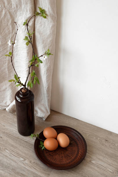 Natural color eggs on brown ceramic plate, vase with green tree branch on table, beige linen curtain, empty white wall with soft sunlight shadows, elegant sustainable rustic Easter kitchen interior.
