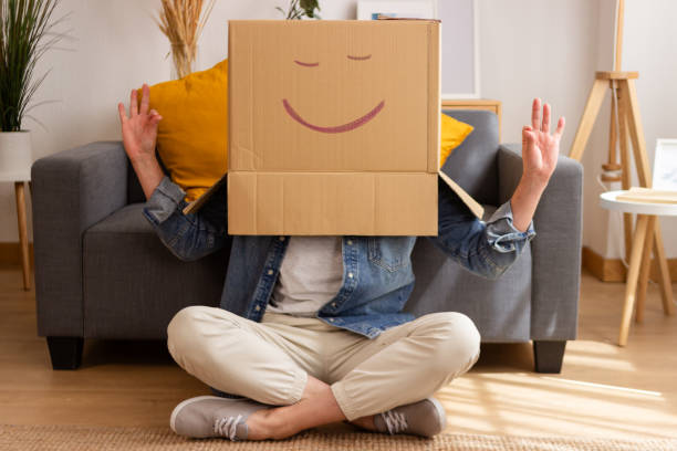 Individual with a cardboard box on their head with a drawn smile sitting cross-legged, radiating positivity