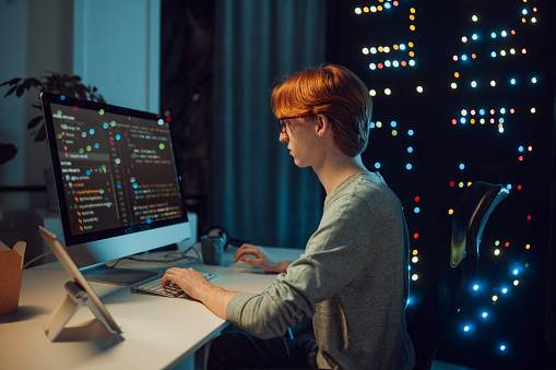 A young red-haired male hacker or programmer is sitting in his workroom, working on a computer. Behind him is a server . He is eating fast food.