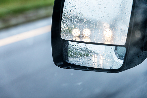 Photo of a speeding delivery van truck's split two section outside rear view mirrors from the driver's perspective/point of view while driving on a highway expressway during a sloppy, torrential downpour rain storm. Two cars with illuminated headlights are visible behind the truck. The one to the left is about to pass the truck. The car appears much smaller and farther away in the lower convex mirror than it does as reflected at 