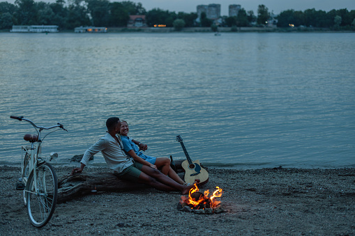 In the evening, a lovely pair is cuddling and chilling by the riverside with a campfire.
