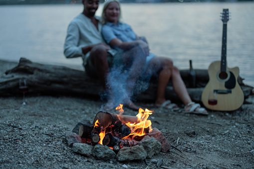 Shot of a bonfire by the river side with a guitar on the side and a couple sitting on a log and hugging