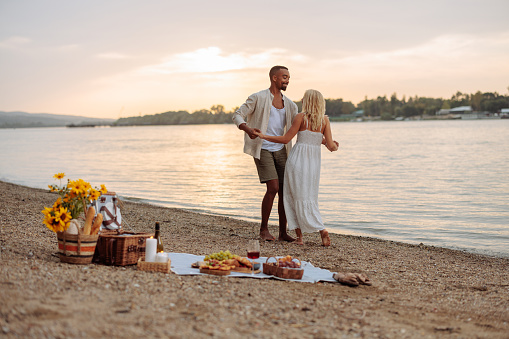 Lovely multiethnic couple  is by the river side, next to them is their picnic blanket with food and drinks, and they are dancing during sunset