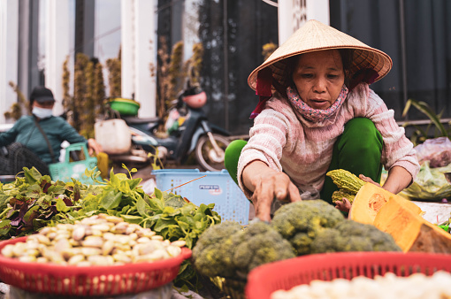 Ho Chi Minh, Vietnam - 4 March 2024 : An old woman selling vegetables at a street market in Ho Chi Minh City. Her traditional hats can be seen across Vietnam