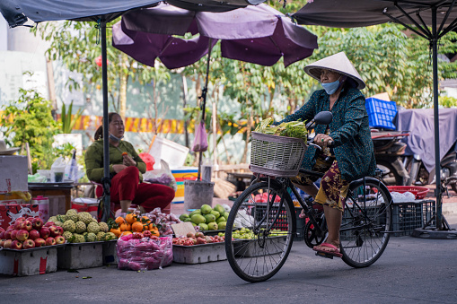 Ho Chi Minh City, Vietnam - March 4, 2024:An old woman riding a bicycle on the street in Ho Chi Minh City, Vietnam, with a street market in the background, Her traditional hats can be seen across Vietnam