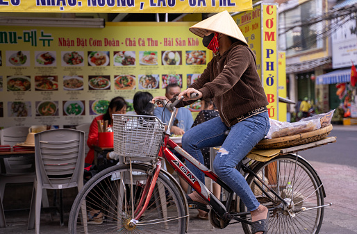 Ho Chi Minh City, Vietnam - March 3, 2024:An old woman riding a bicycle on the street in Ho Chi Minh City, Vietnam, with a street market in the background, Her traditional hats can be seen across Vietnam
