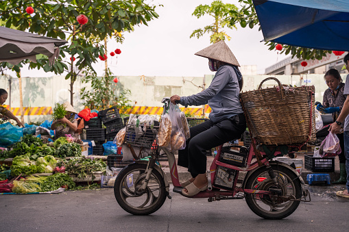 Ho Chi Minh City, Vietnam - March 4, 2024: An old woman riding a motorcycle on the street in Ho Chi Minh City, Vietnam, with a street market in the background, her traditional hat can be seen across Vietnam
