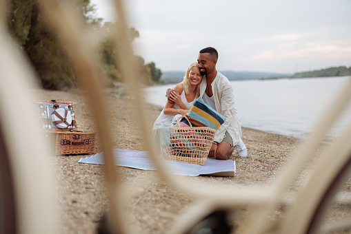 Multiethnic couple is kneeling on a blanket, hugging and smiling before preparing for a picnic by the river
