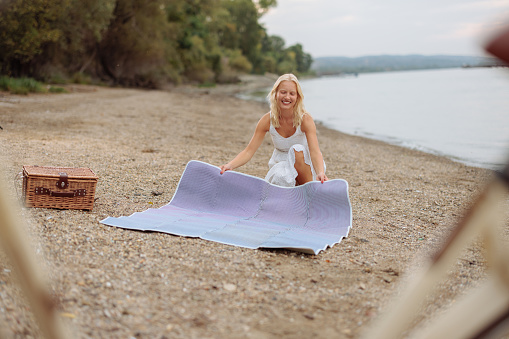 Albino Caucasian girl is spreading her blue blanket on the sand by the river, in order to have a picnic